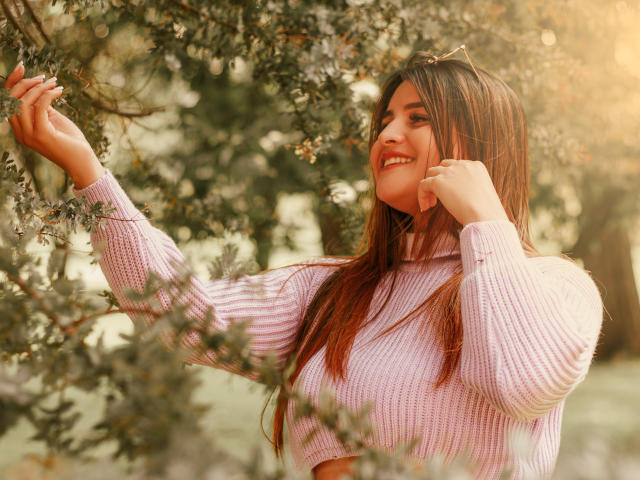 SaraWestt with long brown hair, smiling, wearing a pink knit sweater, touching a tree branch with her right hand, sunlight in her hair, soft green background.