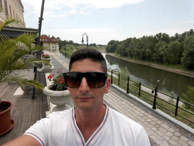 BandiConen69 hat eine niedliche Nase, kurze schwarze Haare und trägt große schwarze Sonnenbrillen zusammen mit einem weißen T-Shirt mit V-Ausschnitt. Er ist draußen in der Nähe eines Flusses, neben einer Pflanze..
