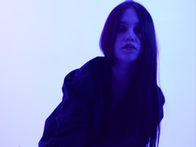 JoannaToy with long dark hair, wearing a black hoodie, has a moody expression, and is bathed in blue light, looking slightly downward. Her tits are small and she's cute.