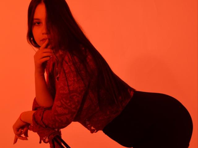 JoannaToy with long dark hair, red lace top, and black pants, leaning forward, finger near mouth, red-lit background, looking seductive, showcasing her ass and sexy pose.