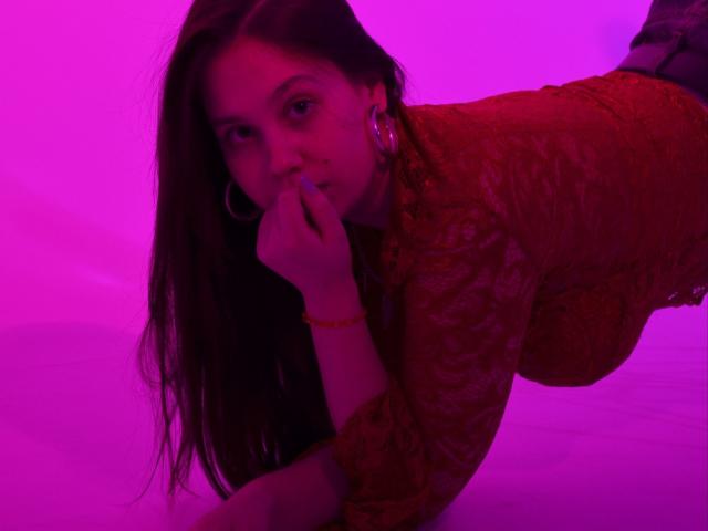 JoannaToy with long brown hair, red lace top, and hoop earrings, lying on her stomach, finger in mouth, pink and red neon lighting, red bracelet on her wrist.