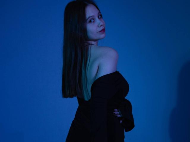 JoannaToy with long brown hair, bare shoulders, black off-shoulder top, looking over her shoulder, blue lighting, soft shadow, sexy, confident, subtle smile.