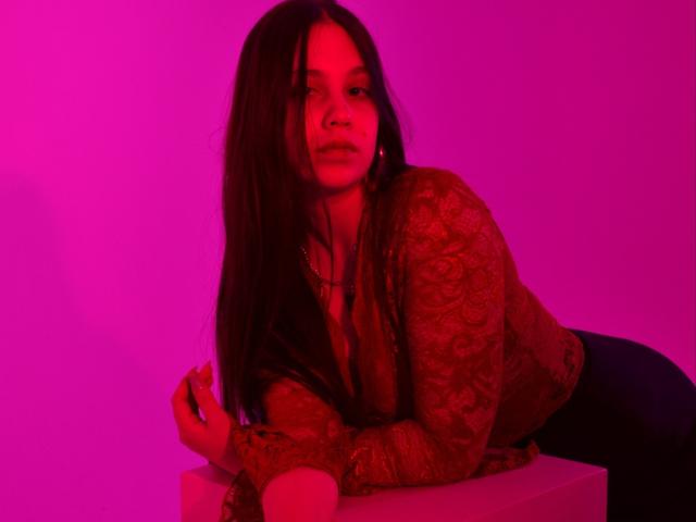 JoannaToy with long dark hair, red-patterned shirt, and dark pants, leaning on a white cube, illuminated by pink-red light, holding a cigarette in her right hand.