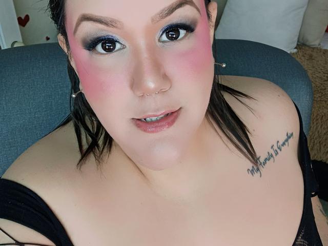 GiiannaMorrys has pink blush, blue eyeshadow, black hair, nose ring, lip gloss, black top, tattoo on right shoulder, looking up with a slight smile, showing her tits.