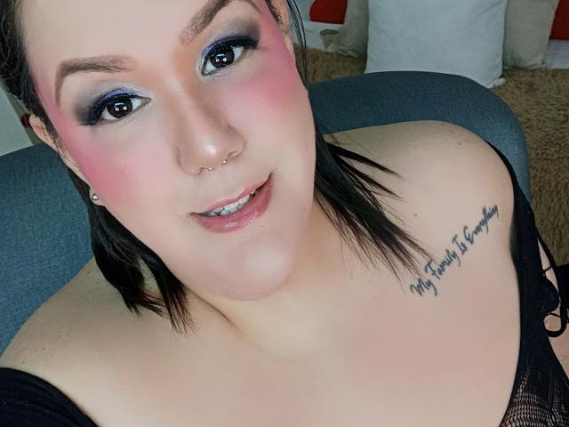 GiiannaMorrys with pink blush, black eyeliner, and nose piercing, smiling at camera, showing bare shoulders and tattoo on right side, wearing black top, has black hair, cute.