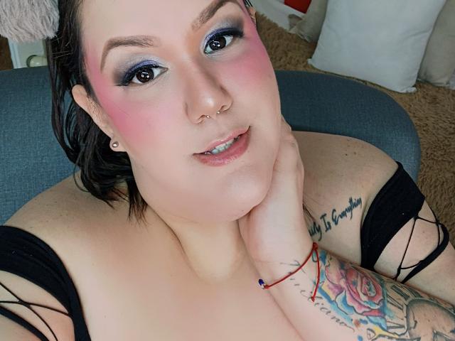 GiiannaMorrys has pink blush on her cheeks, blue eyeshadow, and a nose ring. She's topless with tattoos on her arm and wrist, resting her chin on her hand.