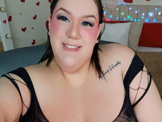 GiiannaMorrys with pink blush, black mesh top, nose ring, and "sassy is forever" tattoo, smiling at camera, showing big tits and chubby ass.