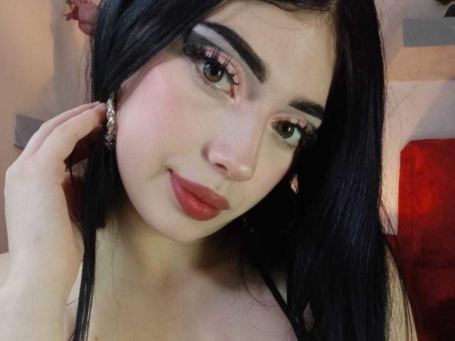 Isabella Rossi heeft lang zwart haar, groene ogen, rode lippenstift en ze raakt haar oor aan met haar hand. Ze draagt roze oogschaduw en een zwarte top..