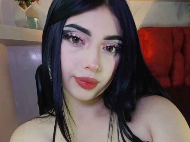 IsabellaaRossi with black hair, pink eyeshadow, long eyelashes, and pink lipstick, showing off her bare shoulders and a nose ring, sitting in a chair.