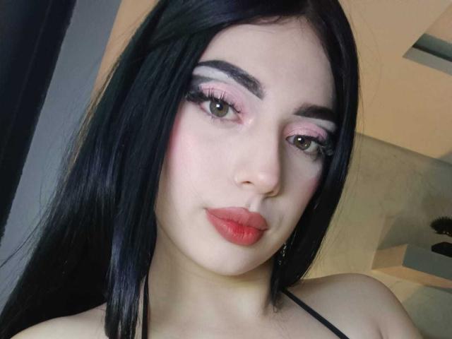 Isabella Rossi, met lang zwart haar, roze oogschaduw, rode lippenstift en volle wenkbrauwen, kijkt in de camera met een lichte glimlach en laat haar blote schouders zien..