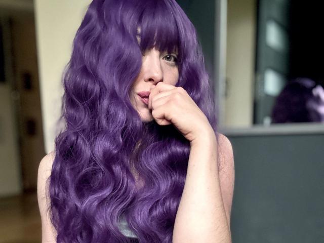 JessieFire-hot with long, wavy purple hair, biting her finger, looking at the camera. She's wearing a green tank top, with a mirror in the background.