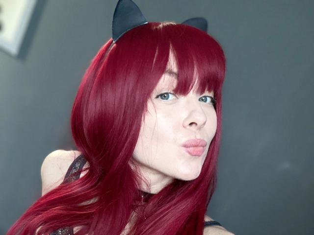 Jessie: Piel ardiente con cabello rojo, orejas de gato negras, labios fruncidos, piel pálida y ojos azules, vestida con una blusa de encaje negro, en un selfie..