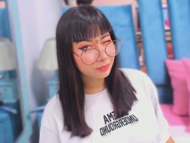 Genesiss has straight black hair, bangs, round glasses, white tee, and a third eye on her forehead. She's winking with pink eyeshadow and a cute smile.