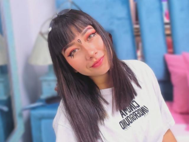 Genesiss with straight black hair, red eye makeup, and a white shirt, showing a cute smile, has a small dot on her forehead, and a cute nose ring.