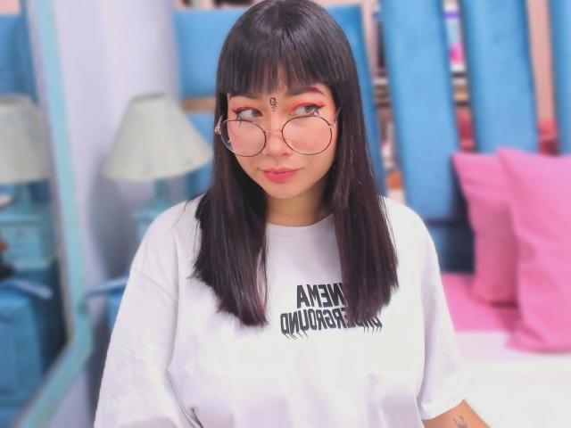 Genesiss with straight black hair, pink-rimmed glasses, white tee, and a forehead bind, showing a cute smile. She's got small tits and a cute, cute face.