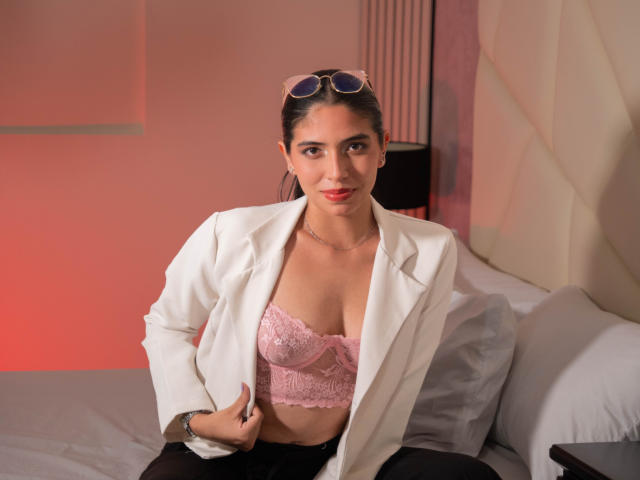 Aria Monroe, con i capelli scuri, indossa un blazer bianco sopra un reggiseno di pizzo rosa, pantaloni neri e occhiali da sole in testa, seduta su un letto, sorridendo..