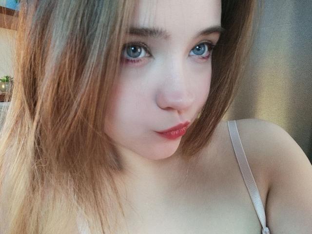CarylPetru has light blue eyes, fair skin, and blonde hair with red lipstick. She's wearing a pink strap, and her small tits are partially visible.