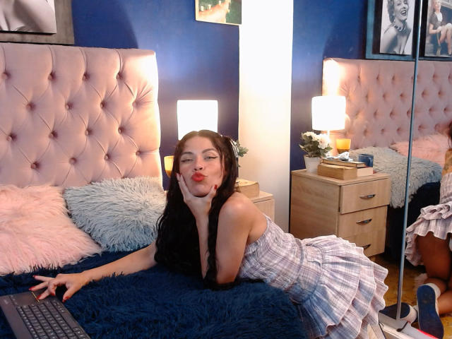 ZoeLux with long black hair, pouty red lips, and a checkered dress, lying on a blue bed, hand on chin, laptop in front, pink headboard, fluff pillows, and wooden nightstand.