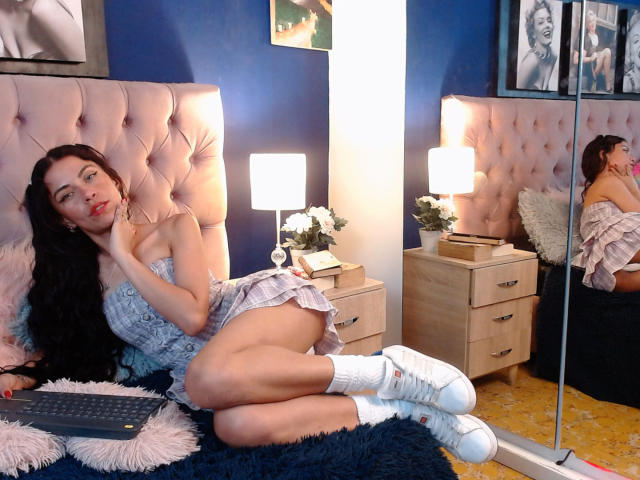 ZoeLux with long black hair, small tits, in a pink tufted headboard bed, wearing a lace-up dress, white socks, and sneakers, lying on a blue rug, laptop beside.