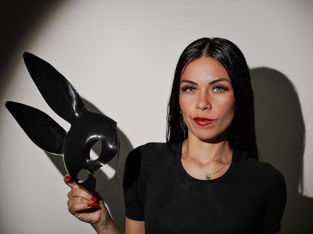 ZoeLux segurando uma máscara preta brilhante da Playboy, com seu longo cabelo preto, batom vermelho e vestido preto. Ela está olhando para a câmera com um leve sorriso, com as unhas pintadas de vermelho..