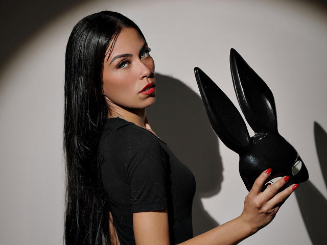 ZoeLux, con lunghi capelli neri, rossetto rosso e camicia nera, che tiene una maschera nera a forma di coniglietta Playboy, unghie rosse, ombra su una parete bianca..