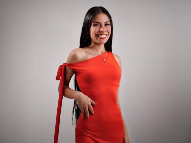 ZoeLux with long black hair, smiling, wearing a tight, off-shoulder red dress with a ribbon, showing her small tits and nipples, standing against a plain gray background.