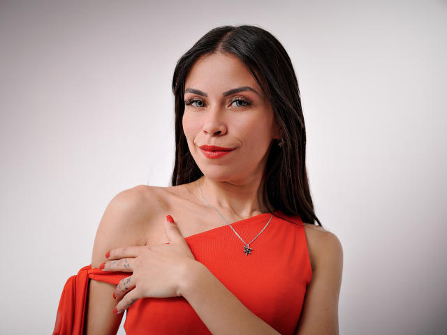 ZoeLux, avec de longs cheveux noirs, une robe rouge et du rouge à lèvres, souriant légèrement, tenant une bretelle de robe, des ongles rouges, un collier avec une petite croix, regardant la caméra..