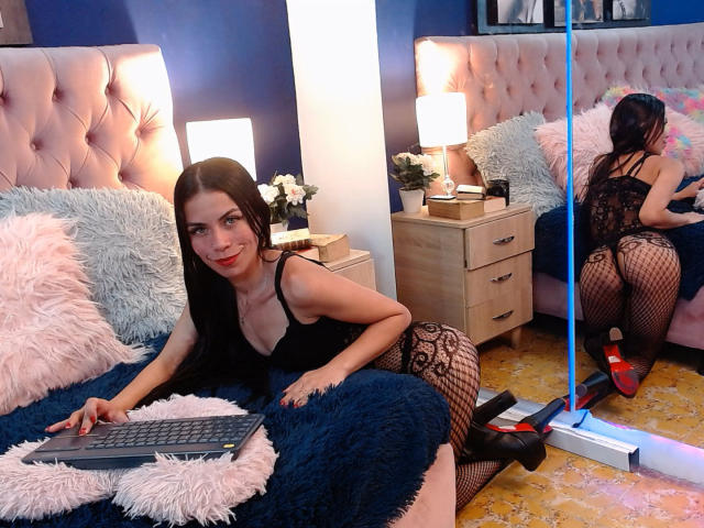 ZoeLux, met zwart haar en rode lippenstift, draagt een zwart kanten body en netpanty's, knielend op een bed, met haar billen naar een spiegel gericht, terwijl ze op een laptop typt, met rode hakken..