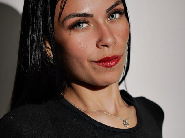 ZoeLux hat ein Nasenpiercing, trägt dunkelroten Lippenstift und hat einen kleinen Muttermal unter ihrem rechten Auge. Sie trägt ein schwarzes Oberteil und eine zarte Halskette. Ihr schwarzes Haar ist glatt..