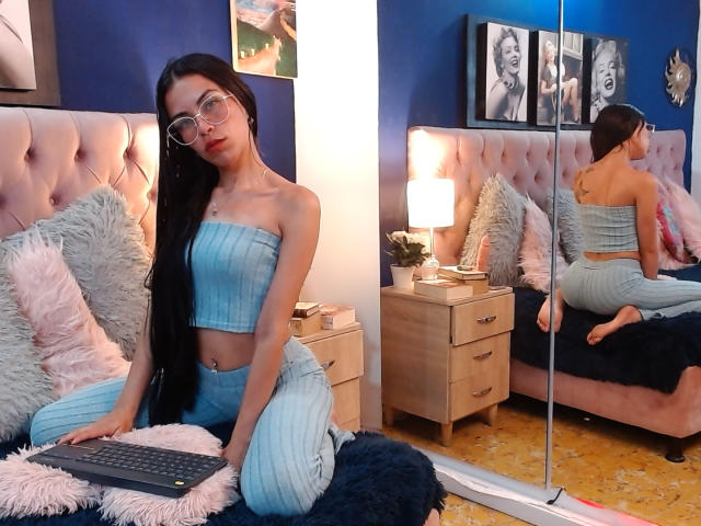 ZoeLux, com cabelo preto longo, óculos, e uma blusa e calça azuis de tecido canelado, ajoelhada em uma cama, refletida em um espelho, com um teclado na frente..