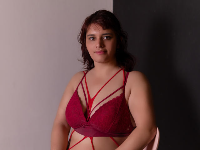 VivianFranco, com pele morena, cabelo castanho e seios pequenos, vestindo um sutiã de renda vermelho com detalhes em tiras, sentada em frente a uma parede preta e branca..