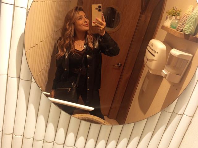 BiancaRey, avec ses cheveux blonds ondulés, sa veste en cuir noir et son pantalon noir, prend un selfie devant un miroir dans une salle de bain, tenant un iPhone doré, et souriant..