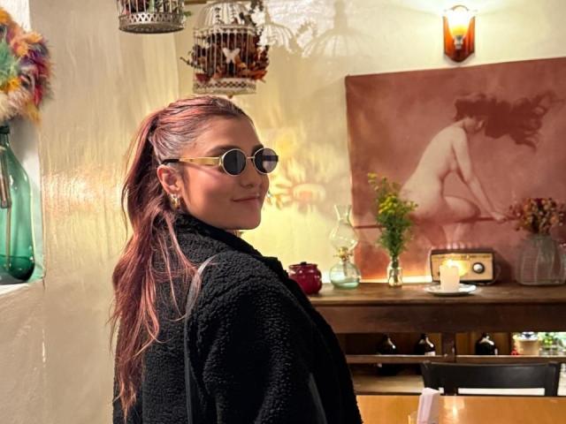 BiancaRey with long pinkish-brown hair in a ponytail, wearing a black textured robe and round sunglasses, smiling over her shoulder, in a warmly lit room.