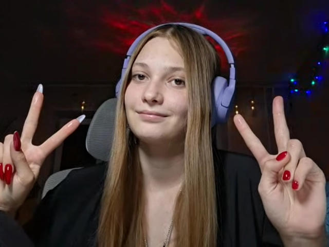 BrittanieManino with long blonde hair, wearing purple headphones, black shirt, red nails, and dual peace signs, smiling slightly, in a dimly lit room with colorful lights.