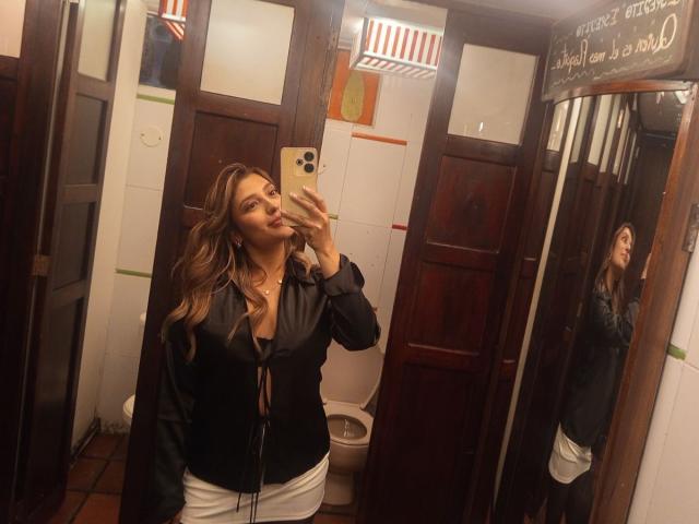 BiancaRey, con i capelli castano ondulati, una maglietta nera e pantaloni bianchi, sta scattando un selfie allo specchio, mostrando il seno e i glutei in un bagno di legno, mentre tiene in mano un iPhone dorato..