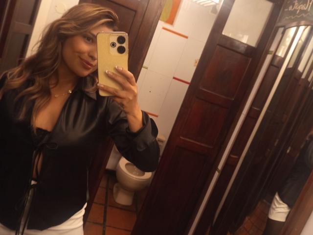 BiancaRey with wavy brown hair, smiling, taking a mirror selfie in a bathroom, wearing a black leather jacket, holding a gold iPhone, showing her tits and ass.