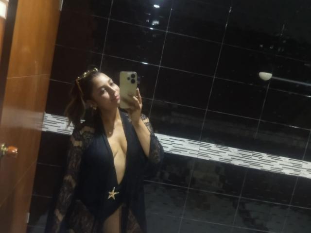 BiancaRey with big tits, wearing a black lace robe, open to show cleavage, holding a phone in a mirror selfie, dark tile background, looking at the camera.