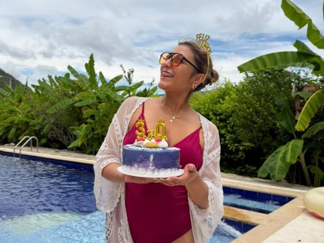 BiancaRey stands by a pool, wearing a red one-piece, sheer white cover-up, and glasses, holding a blue birthday cake with a "4" candle, smiling. Her tits are visible.
