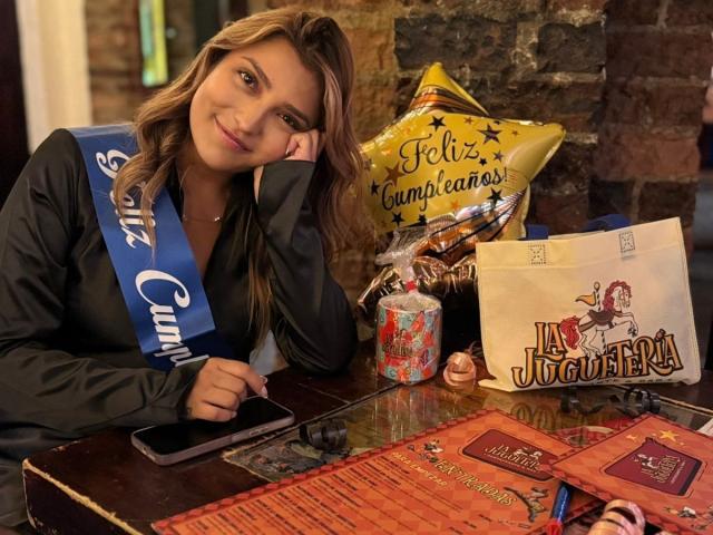 BiancaRey with brown hair, wearing a blue sash, black shirt, and holding a phone, next to snacks, a bag, and a red mat on a wooden table.