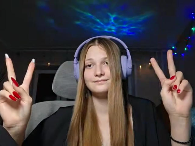 BrittanieManino with long blonde hair, red nails, and white headphones, making double peace signs, wearing a black shirt, in a dimly lit room with blue ceiling lights.