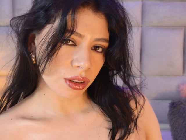 Linda Mancini ha i capelli scuri e ondulati, labbra piene con un rossetto rosa lucido, e indossa piccoli orecchini dorati. La sua pelle è chiara, con una texture liscia e leggermente lucida..