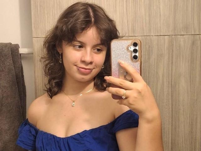 GagingMolly with curly brown hair, smiling, holding phone, wearing off-shoulder blue top, showing small tits, light skin, necklace, ring, and brown towel in background.