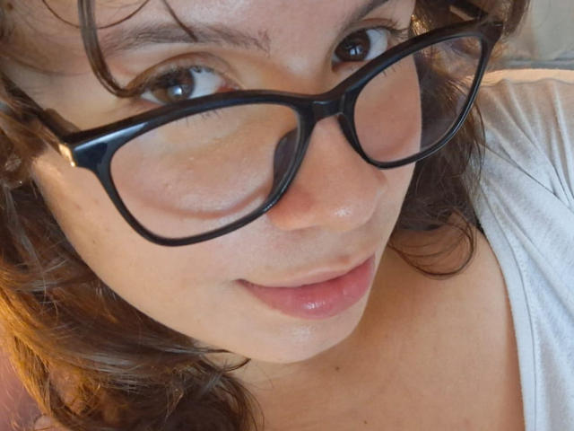 GagingMolly with glasses, brown curly hair, light skin, and pink lips, wearing a white shirt, showing a slight smile, her face centered in the shot.