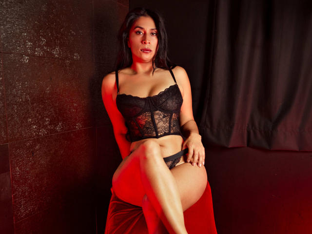 AfroditaDoir, con cabello oscuro, vestida con un sujetador y tanga de encaje negro, sentada sobre un paño rojo, exponiendo su vulva, con las piernas cruzadas, mirando seductoramente a la cámara..