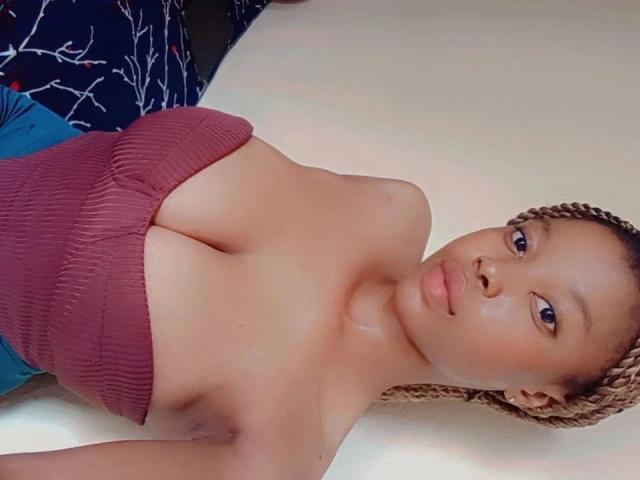 Sexypussy-hot with braided hair, lying on a white surface, wearing a strapless, ribbed, red top that shows her big, shiny tits and a slight smile on her face.