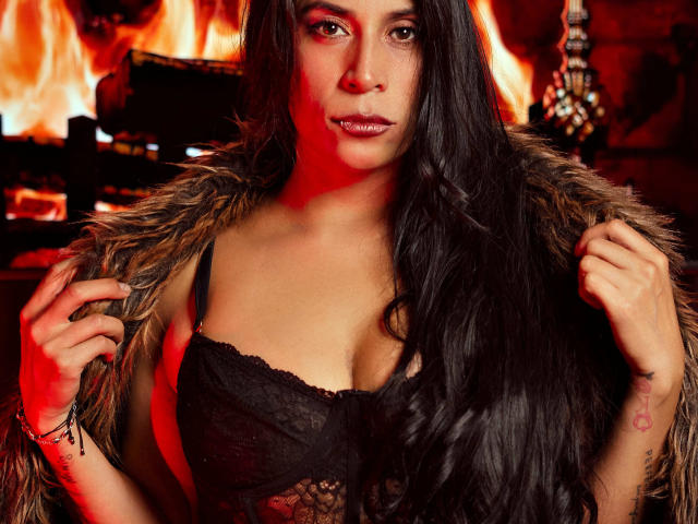 AfroditaDoir, avec de longs cheveux noirs, portant un soutien-gorge noir en dentelle et un manteau en fourrure, qu'elle tient ouverte, dévoilant ses seins, debout devant un feu, regardant l'appareil photo..