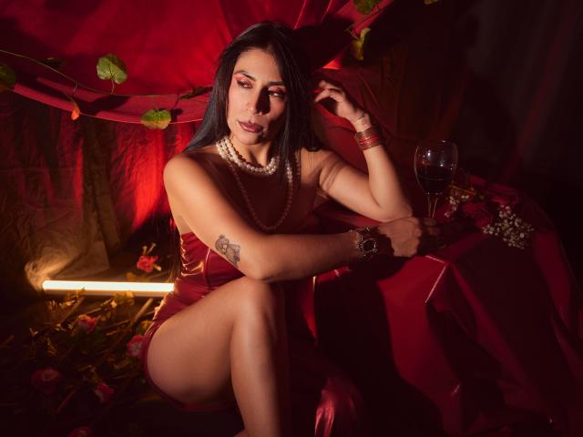AfroditaDoir con cabello oscuro, vestido rojo, collar de perlas, sentada sobre una tela roja, con las piernas cruzadas, sosteniendo una copa de vino, con un tatuaje en el brazo izquierdo, con una mirada seductora, iluminación roja tenue..