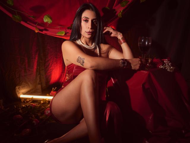 AfroditaDoir con cabello oscuro, corpiño rojo, collar de perlas, tatuaje en el brazo derecho, sentada con las piernas cruzadas, una mesa roja, una copa de vino, un paño rojo y hojas, todo iluminado con una luz roja tenue..