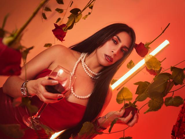 AfroditaDoir, avec de longs cheveux noirs, une robe rouge, un collier de perles, tenant un verre de vin rouge, entourée de roses et de feuilles vertes, sous une lumière rouge, regardant l'appareil photo..