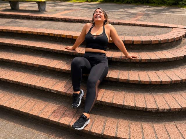 EmaFoxy está sentada en unos escalones de ladrillo, riendo con una expresión de alegría. Lleva un top deportivo negro, leggings negros y zapatillas negras, y tiene las manos apoyadas en los escalones..
